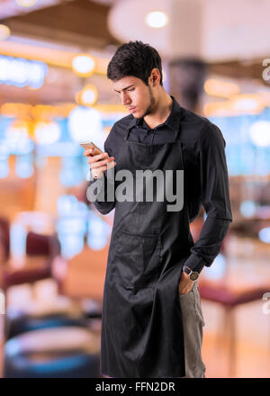 Jeune cuisinier ou serveur posant, le port de tablier noir et chemise blanche, à l'aide de téléphone cellulaire ou votre téléphone intelligent ou dispositif d'ordres, en bar, r Banque D'Images