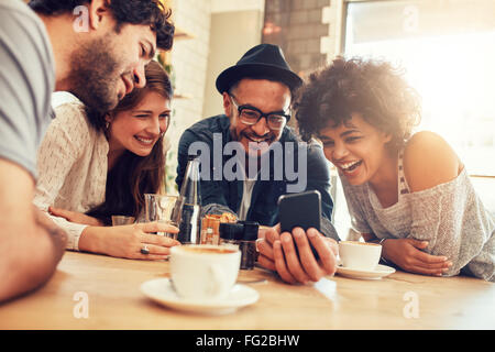 Portrait of cheerful young friends looking at smart phone while sitting in cafe. Mixed Race des gens assis à une table de restaurant Banque D'Images