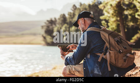 Man avec sac à dos en utilisant l'onglet numérique tout en étant assis près d'un lac. Homme mûr sur la randonnée dans la nature à l'aide de tablette numérique. Banque D'Images