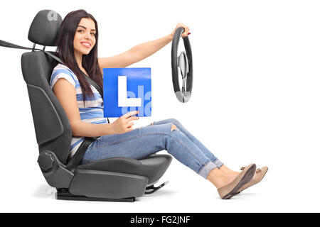 Jeune femme tenant un L, signer et un volant assis sur un siège de voiture isolé sur fond blanc Banque D'Images