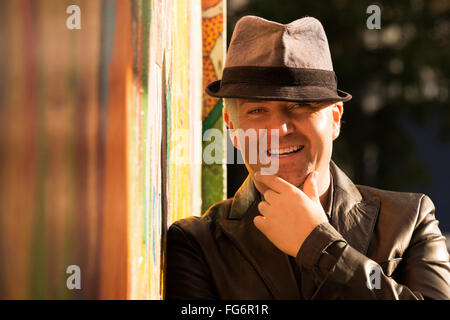 Portrait of a smiling man dans un manteau de cuir et chapeau gris avec sa main sur son menton, debout contre un mur ensoleillé avec une murale colorée sur elle Banque D'Images