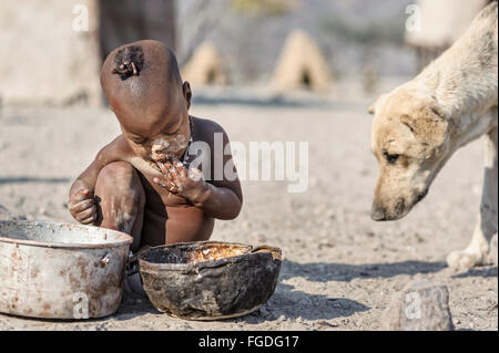 Enfant Himba assis sur le sol de son village éloigné de manger un pot rouillé tandis qu'un chien est à proximité de lui. Banque D'Images