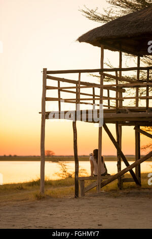 Une jeune femme est assise sous un tourisme hutte au toit de chaume et prend des photos de coucher de soleil tout en safari dans le Makgadikgadi Pans, Botsw Banque D'Images