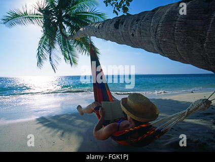 Une femme dans un hamac en lisant un livre sous un cocotier tree refoulées au fil de l'eau sur une plage sur la côte Pacifique du Costa Rica Banque D'Images