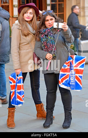 Les jeunes femmes chaque exploitation union jack panier préparation à l'utilisation d'un stick & selfies téléphone d'Apple tout en shopping à Londres Piccadilly Circus Banque D'Images