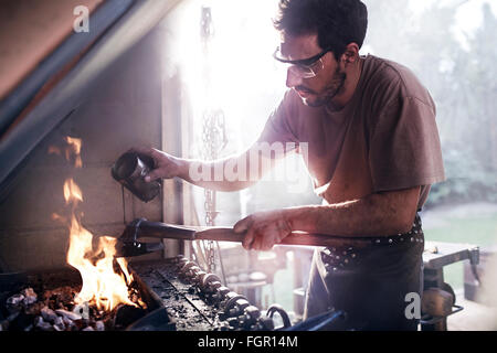 Blacksmith pouring liquid sur l'outil en feu en forge Banque D'Images