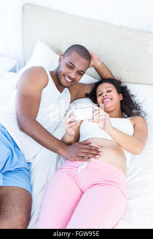 Pregnant wife holding smartphone avec mari lying on bed Banque D'Images