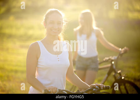 Deux belles copines cyclistes occasionnels porter white tops et short jeans debout avec des vélos dans le parc de campagne Banque D'Images