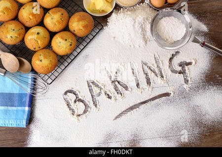 Le mot écrit dans la farine à pâtisserie sur une table en bois sombre avec des muffins et des ingrédients. Banque D'Images