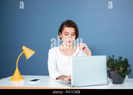 Concentré pensif jeune femme assise à la table de travail avec ordinateur portable et sur fond bleu Banque D'Images