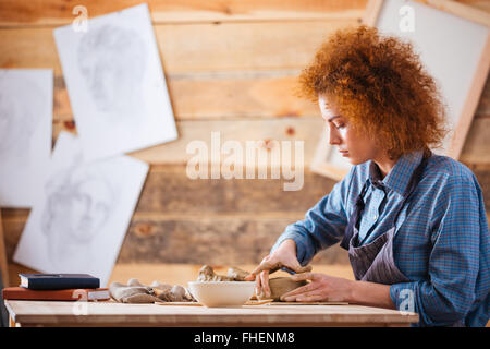 Jeune femme rousse sérieux potter assis et créant des plats avec de l'argile par des mains Banque D'Images