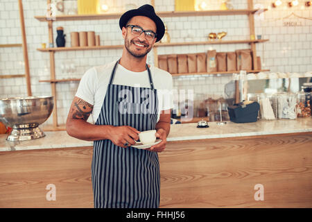 Portrait of happy young barista au travail. Caucasian man wearing apron et hat standing in front of cafe comptoir avec tasse de café Banque D'Images