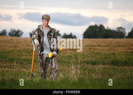 Chasseur de trésor avec détecteur de métal s'appuya sur une pelle dans le champ. Vêtements de camouflage et d'oeil confiant. Contexte Rural Banque D'Images