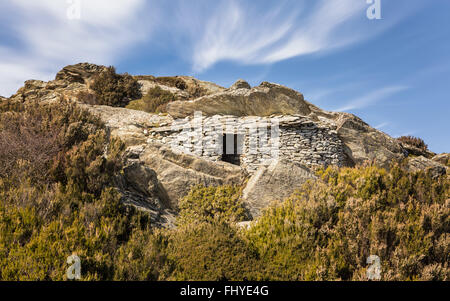 Un abri en pierre construit dans la roche entre le maquis dans les montagnes du nord de la Corse, avec un ciel bleu et des nuages filandreux Banque D'Images
