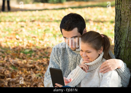 Young couple in autumn park assis près de la lecture de l'arbre du livre électronique Banque D'Images
