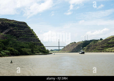 Pont Centennial Canal de Panama Ciudad de Panama Panama | CANAL DE PANAMA, Panama — Un remorqueur passe dans la Coupe Gaillard sous la distante Pont Centennial, dont le pylône unique s’élève entre les berges creusées qui marquent le passage le plus étroit du Canal de Panama. Achevée en 2004 pour le centième anniversaire de la République de Panama, le pont à haubois offre une hauteur de libération verticale de 80 mètres (262 pieds) pour les navires Panamax et transporte l’autoroute panaméricaine à travers le canal près des écluses de Pedro Miguel. Banque D'Images