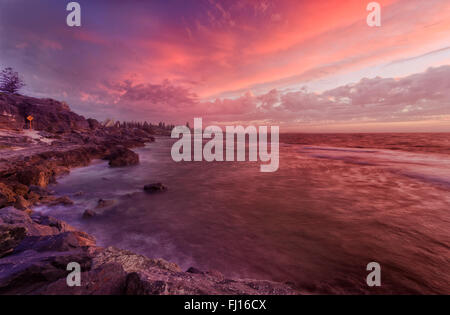 Face à l'ouest majestueux coucher du soleil à Perth Cottlesloe beach. Rochers dangereux attaqué par floue ondes sous ciel nuageux. Banque D'Images