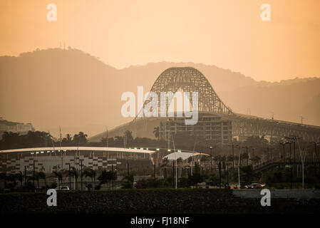 Pont des Amériques Canal de Panama Panama City | PANAMA CITY — La brume du soleil couchant encadre le Pont des Amériques enjambant le Canal de Panama, avec le complexe sportif Maracaná visible au premier plan de Panama City. Achevé en 1962, le pont à arche en acier porte l’autoroute panaméricaine à environ 384 pieds (117 m) au-dessus de l’eau et était le seul passage fixe du canal jusqu’à l’ouverture du Pont du Centenaire en 2004. Banque D'Images
