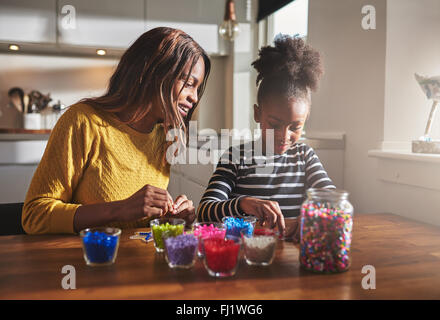 Femme heureuse et jeune fille assise et la création d'artisanat de perles avec différentes couleurs sur table en bois dans la cuisine avec du soleil comin Banque D'Images