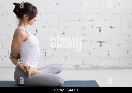Belle jeune femme travaillant à l'intérieur, faisant de l'ouvreur de la hanche, yoga exercice sur tapis bleu, assis en Padmasana Baddha, lié Banque D'Images