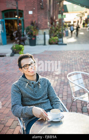 Young Asian man sitting and smiling in outdoor cafe holding relaxant tasse de café, avec copie espace Banque D'Images