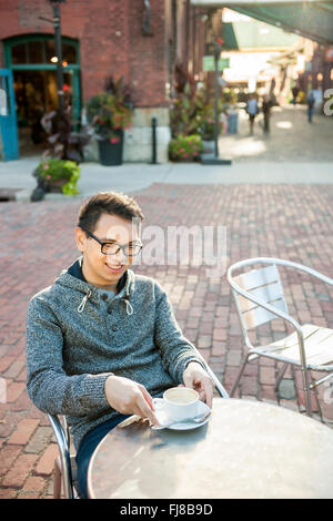 Young Asian man sitting and smiling in outdoor cafe holding relaxant tasse de café à la bas Banque D'Images