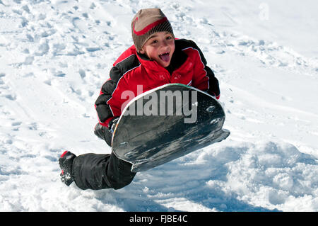 Cute boy dans la descente de luge une luge sur une colline de neige avec la langue qui sort. Banque D'Images