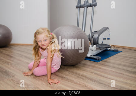 Petite fille blonde dans la salle de sport avec des équipements de sport et de balle gris assis sur le plancher Banque D'Images