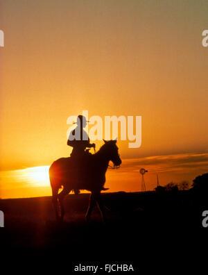 AMERICAN COWBOY, Cowboy riding son cheval au coucher du Soleil avec ferme et moulin en arrière-plan. New York, USA Banque D'Images