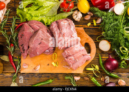 Tranches de morceaux de viande rouge sur une planche à découper plus de grain du bois table avec divers légumes verts et herbes qui l'entourent Banque D'Images