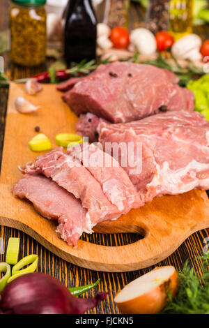 Des morceaux de viande rouge sur une planche à découper en tranches avec les oignons dans l'avant-plan et d'autres légumes et d'ingrédients à l'arrière-plan. Banque D'Images