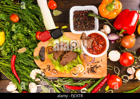 Angle de vue de la vie toujours élevé de petits morceaux de boeuf sur une planche à découper en bois avec bol de trempette Salsa entouré d'abondance de produits frais Banque D'Images