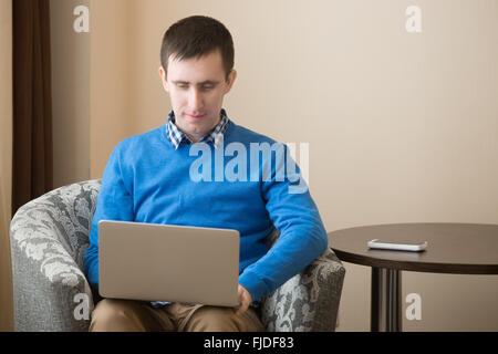 Portrait of attractive young casual homme assis dans un fauteuil à l'intérieur, à l'aide d'un ordinateur portable, regarder l'écran, male Banque D'Images