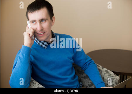 Portrait of attractive occasionnels détendus smiling young man sitting in armchair intérieur, holding cellphone Banque D'Images