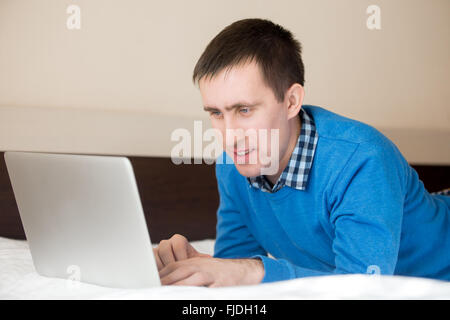 Portrait of smiling casual young man lying on bed using laptop, travailler, modèle masculin à la recherche à l'écran Banque D'Images