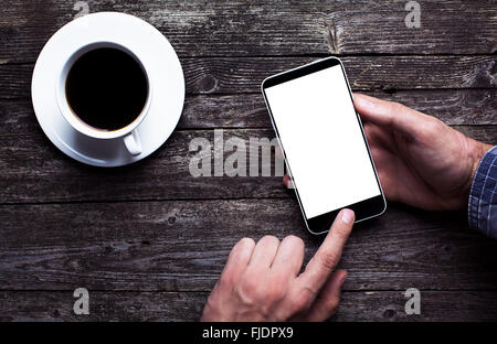 Dans les mains d'hommes chemise en utilisant smartphone avec grand écran tactile blanc sur noir en bois rustique avec une tasse de café. Avobe voir télévision lay Banque D'Images