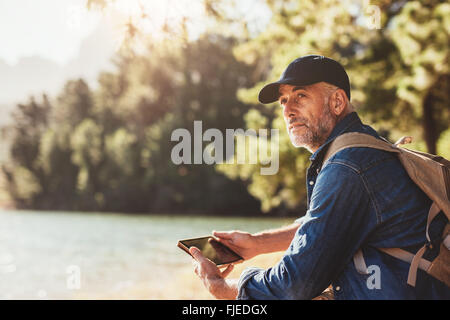 Portrait of senior homme assis près d'un lac avec sac à dos et tablette numérique. Young male hiker assis à un lac et à la awa Banque D'Images