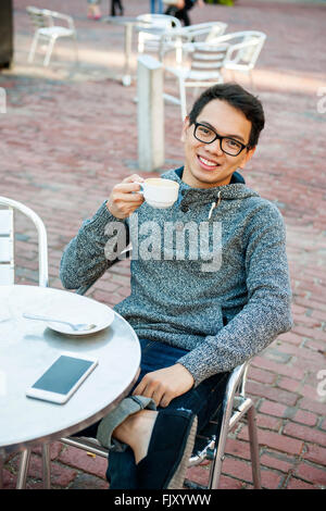 Young Asian man sitting and smiling in cafe de détente en plein air avec un téléphone mobile holding tasse de café Banque D'Images