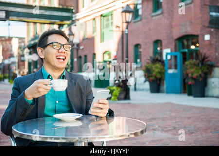Laughing young asian man sitting in outdoor cafe with mobile phone holding tasse de café du succès Banque D'Images