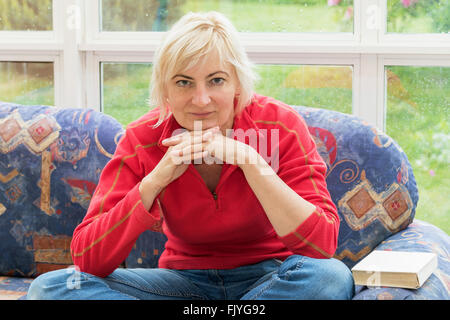 Femme blonde est assise sur le canapé et est à la recherche de l'appareil photo. Femme est étaiement menton avec ses mains. Banque D'Images