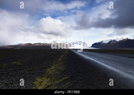 Photo horizontale d'une route asphaltée tout droit venant de la montagne avec les nuages au-dessus et le glacier de Vatnajokull dans le backg Banque D'Images