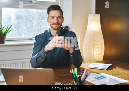 Jeune entrepreneur assis à son bureau avec une tasse de café en souriant à l'appareil photo Banque D'Images