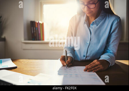 Seule la femme en bleu de travail blouse à 24 aux formalités administratives en home office avec la lumière du soleil sur son épaule à travers la vitre Banque D'Images