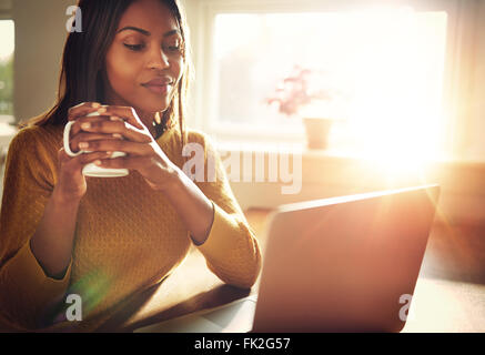 Hot woman smiling sitting près de fenêtre lumineuse tout en regardant l'ordinateur portable ouvert sur la table et holding white mug Banque D'Images