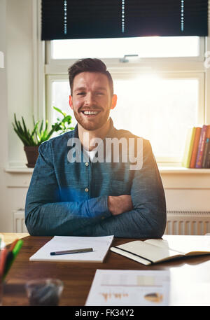 Smiling happy homme assis à son bureau avec les bras pliés de rire à l'appareil photo avec la paperasse sur le bureau en f Banque D'Images