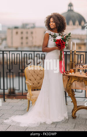 Charmante fiancée noire avec bouquet de mariage en mains debout sur la terrasse Banque D'Images