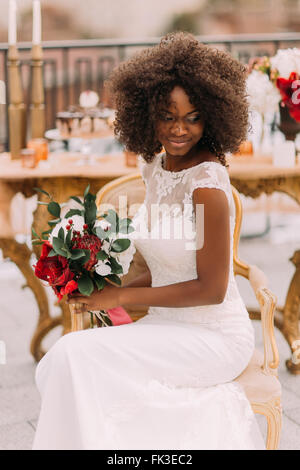 Heureusement mariée noir merveilleux sourire avec les yeux fermés et tenant un bouquet de fleurs rouges. Jour de mariage Banque D'Images