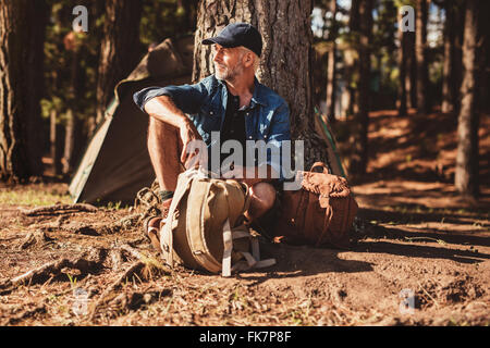 Portrait of senior homme assis sous un arbre avec un sac à dos. Homme mûr assis seul au camping en forêt. Banque D'Images