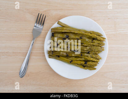 Vue de dessus d'une portion d'asperges grillées trempées dans l'huile de tournesol sur une plaque blanche avec une fourchette pour le côté au-dessus d'une table en bois Banque D'Images