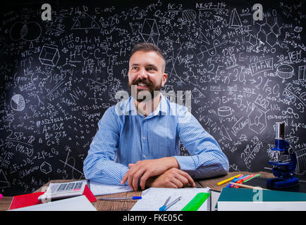 Happy teacher sitting at desk, fournitures scolaires, grand tableau noir Banque D'Images